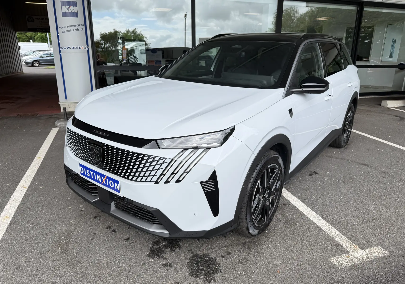Peugeot 5008 HYBRID 145 e-DCS6 GT blanc Okenite avec toit noir, vue 3/4 avant droit sur parking devant vitrine.