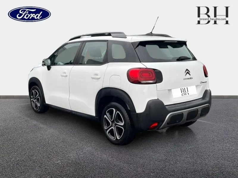 Vue 3/4 arrière droite d'un Citroën C3 Aircross blanc Natural White avec barres de toit et feux arrière distinctifs.