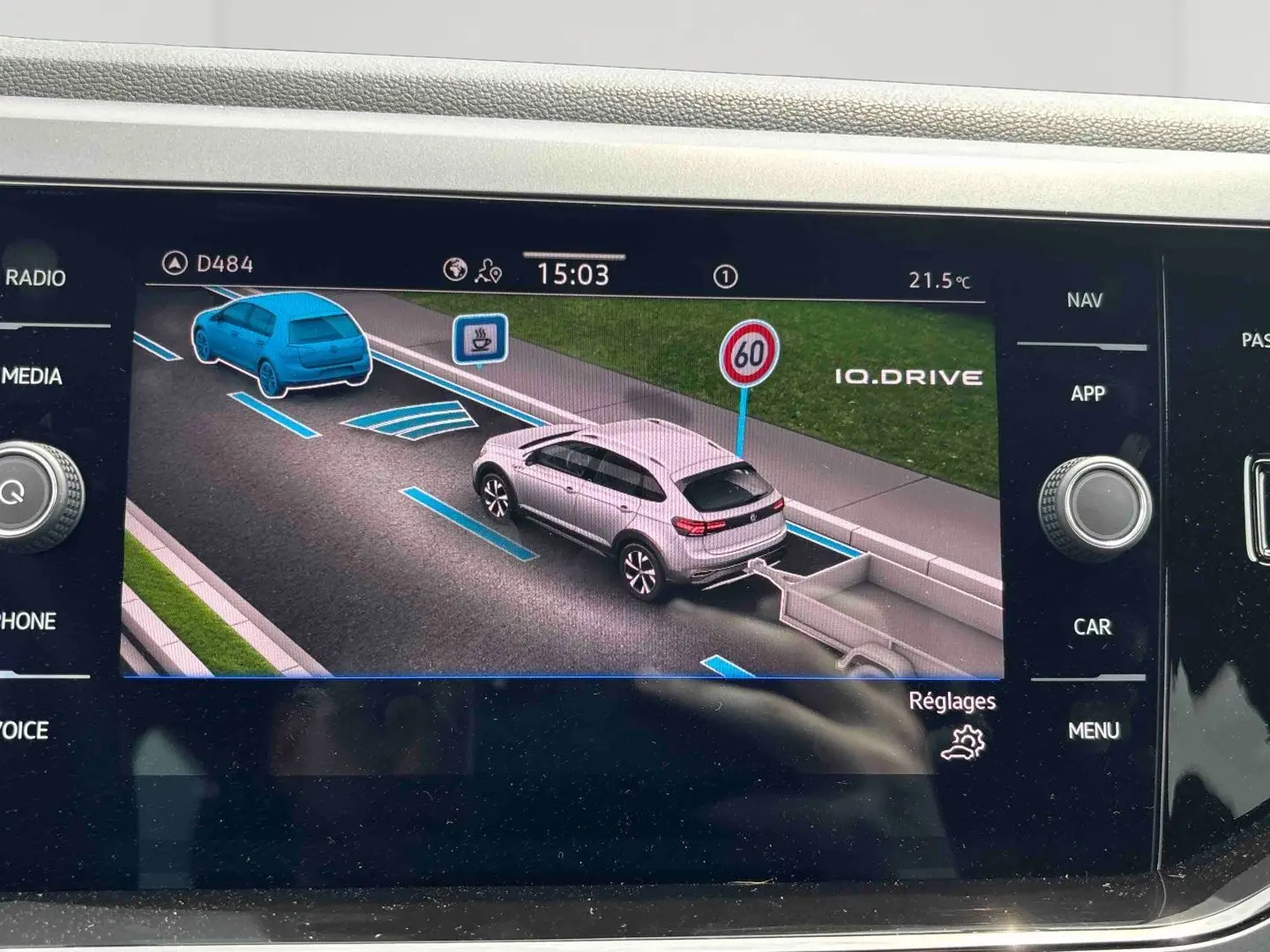 Écran tactile montrant une vue 3/4 arrière d’un Volkswagen Taigo blanc avec attelage sur route et panneau 60 km/h.