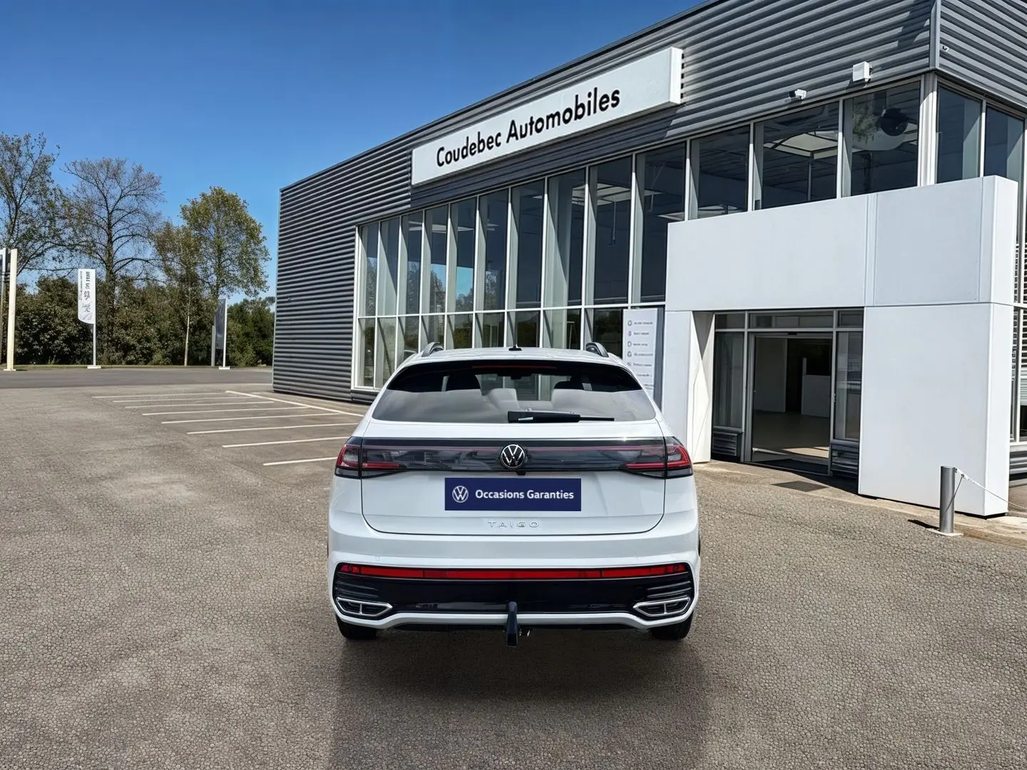 Vue arrière d’un Volkswagen Taigo blanc 2025 avec attelage amovible, garé devant un bâtiment de concession.
