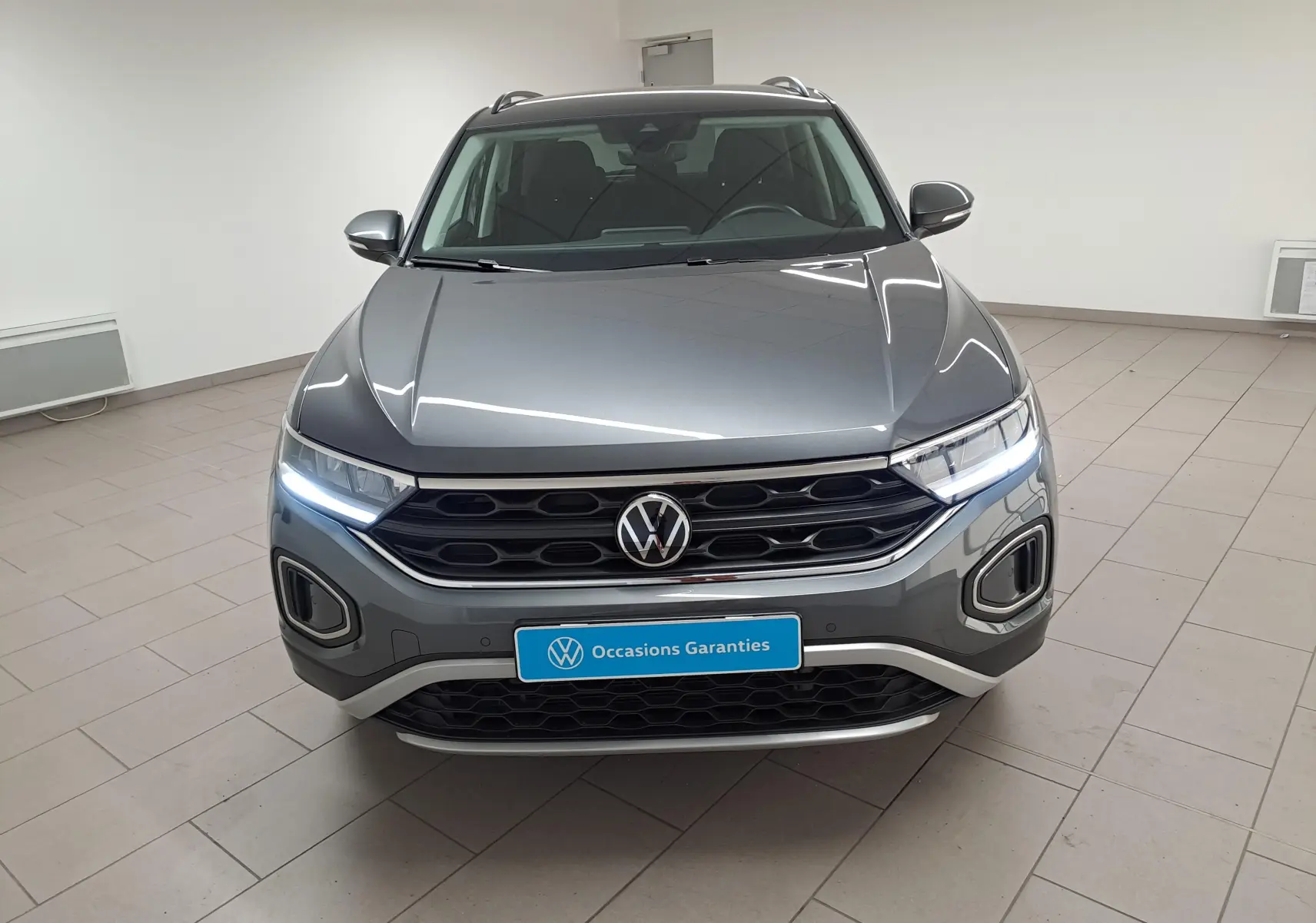 Vue frontale d'un Volkswagen T-Roc 2024 gris Indium avec phares allumés et calandre noire distinctive.