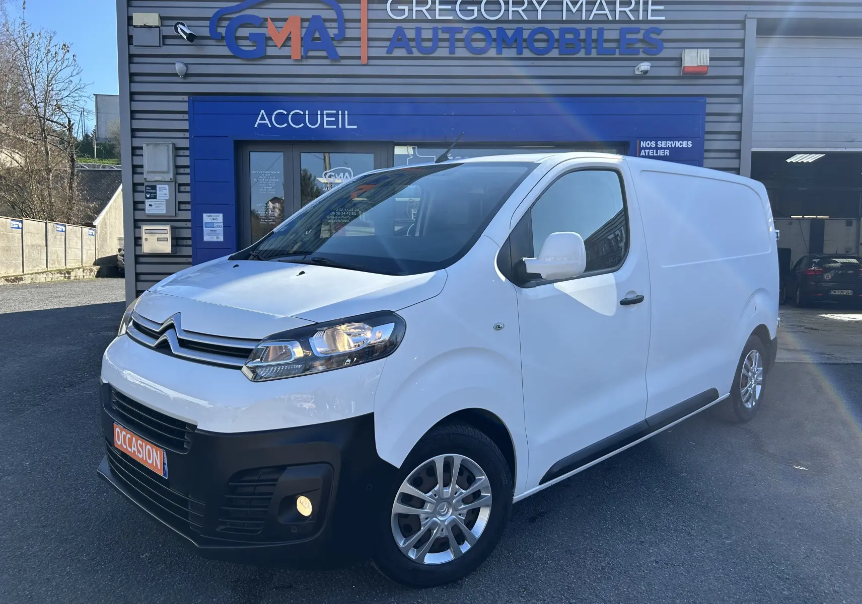 Vue 3/4 avant droite d'un Citroën Jumpy blanc avec attelage, garé devant un bâtiment commercial.