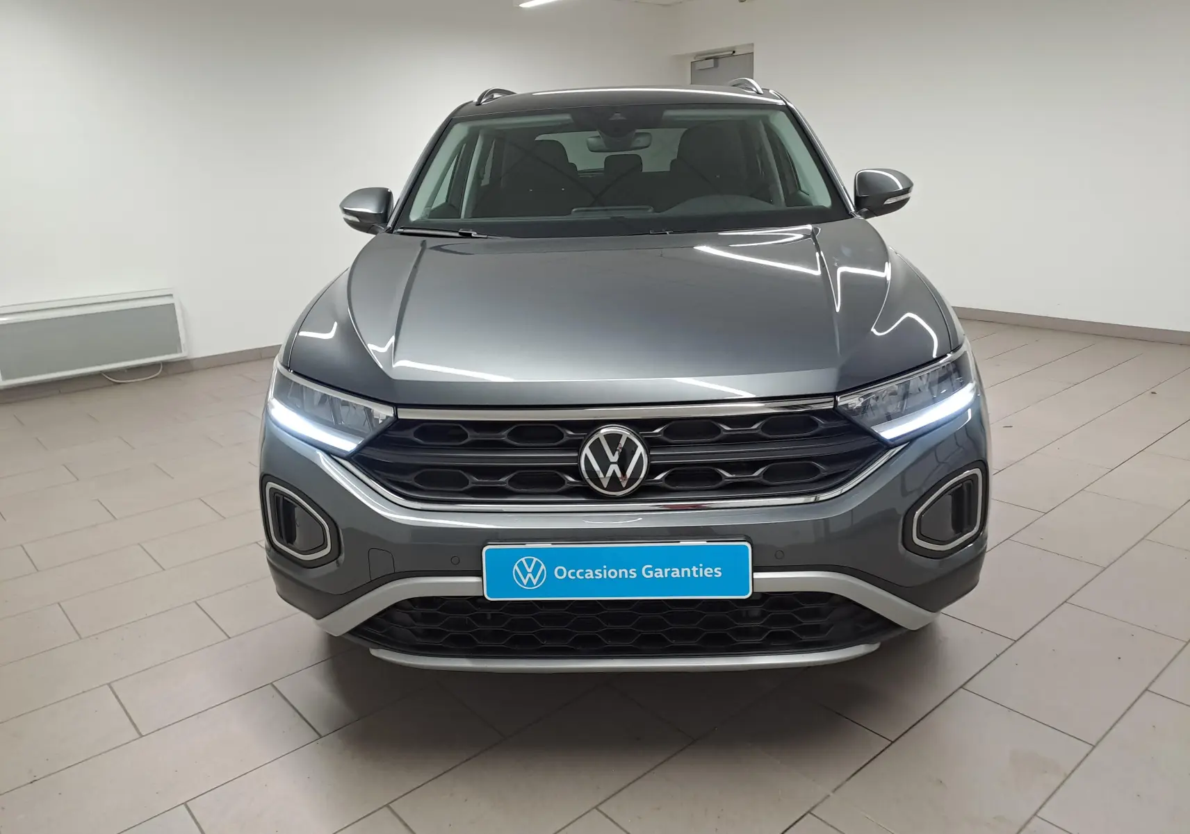 Vue frontale d'un Volkswagen T-Roc gris Indium avec feux LED allumés et calandre noire dans un intérieur neutre.