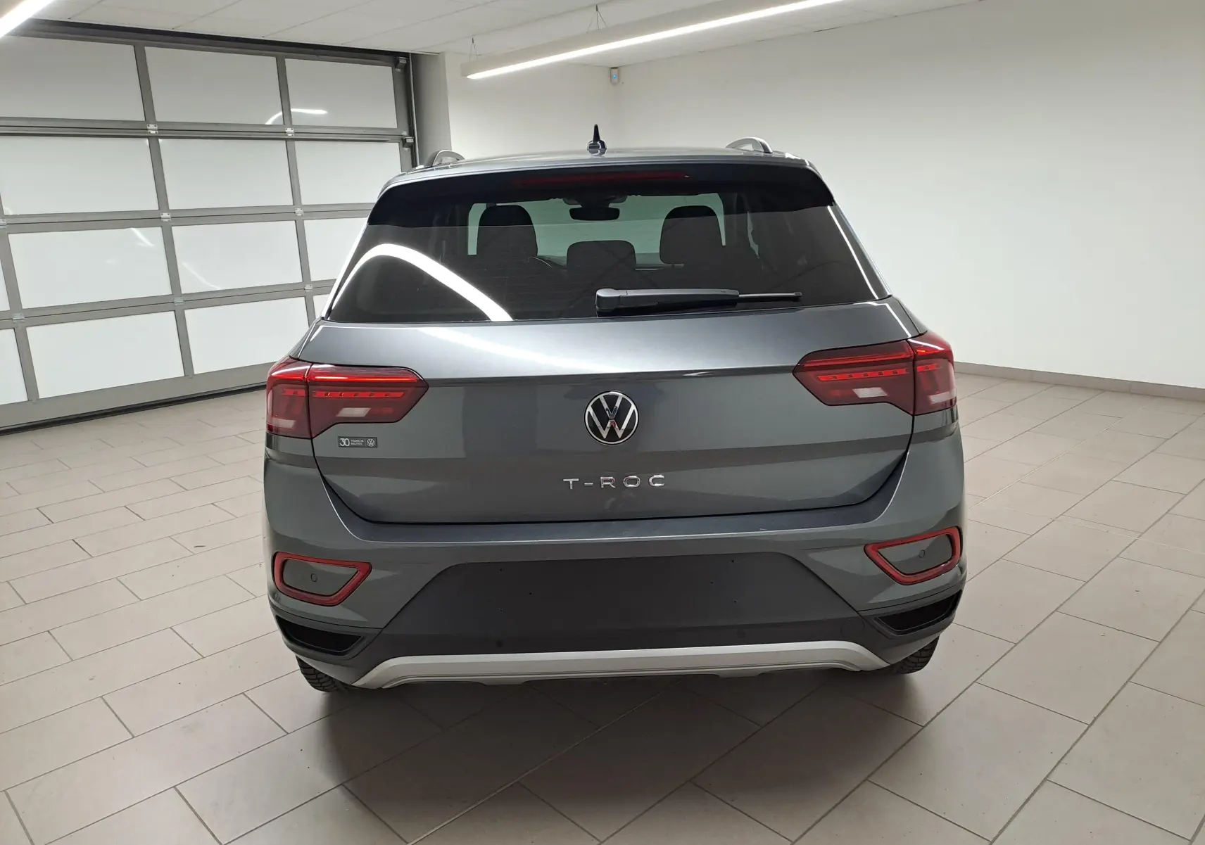 Vue arrière d'un Volkswagen T-Roc 2024 gris Indium avec feux arrière allumés et caméra de recul visible en intérieur neutre.