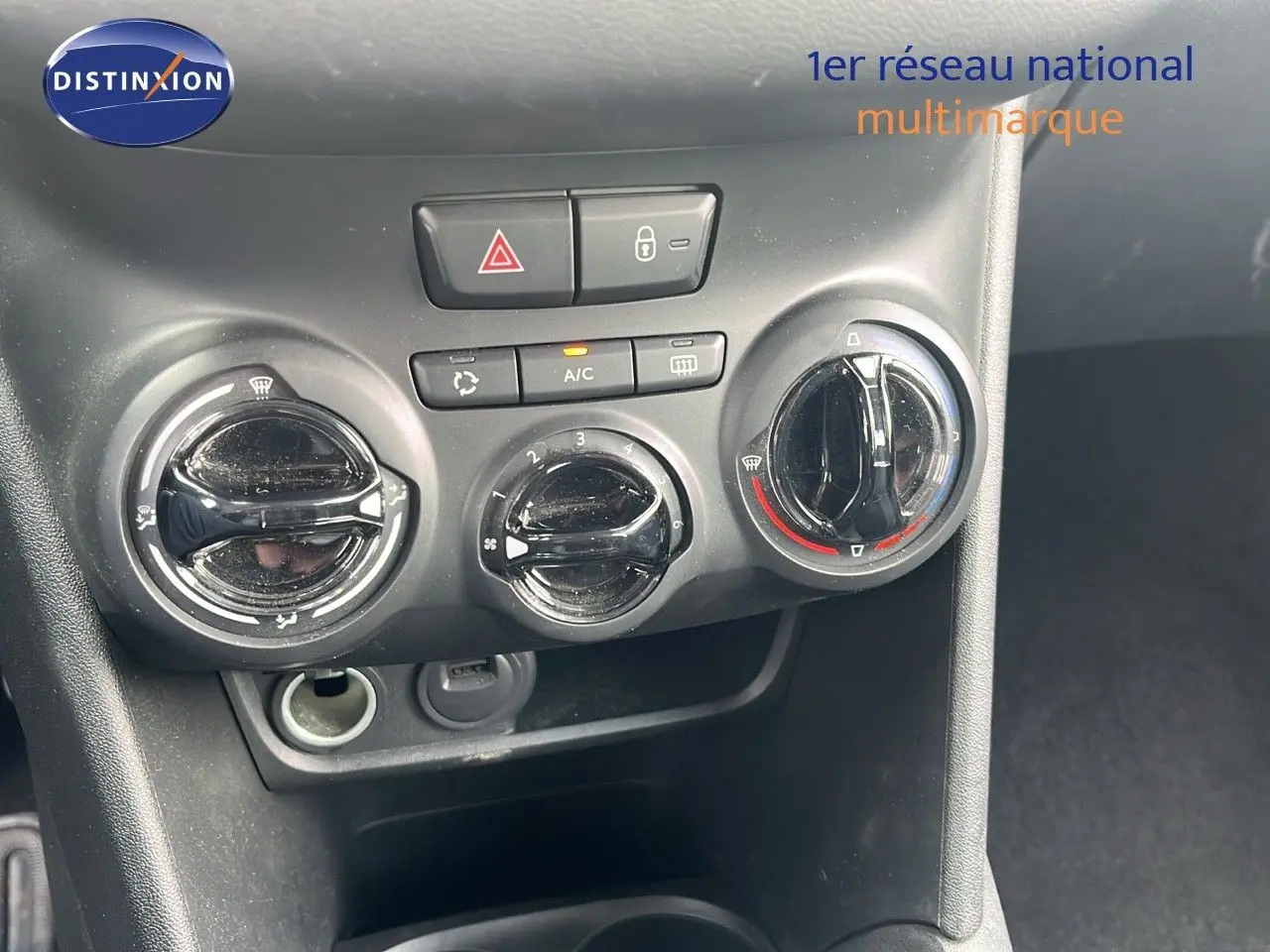 Gros plan sur les commandes de climatisation et les boutons de sécurité du tableau de bord d'une Peugeot 208 blanche.