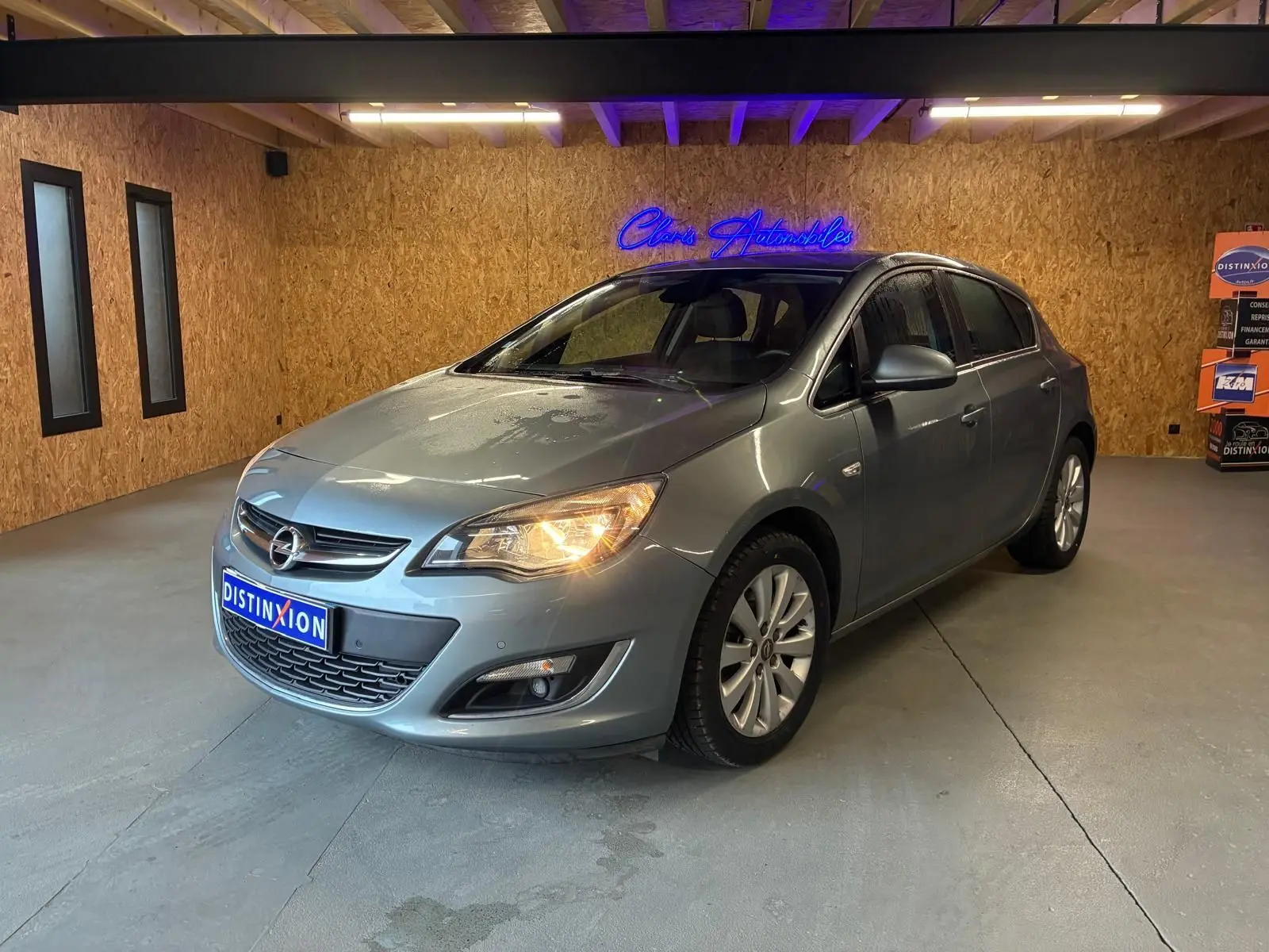 Opel Astra gris 2015 vue 3/4 avant droit avec phares allumés dans un showroom intérieur.