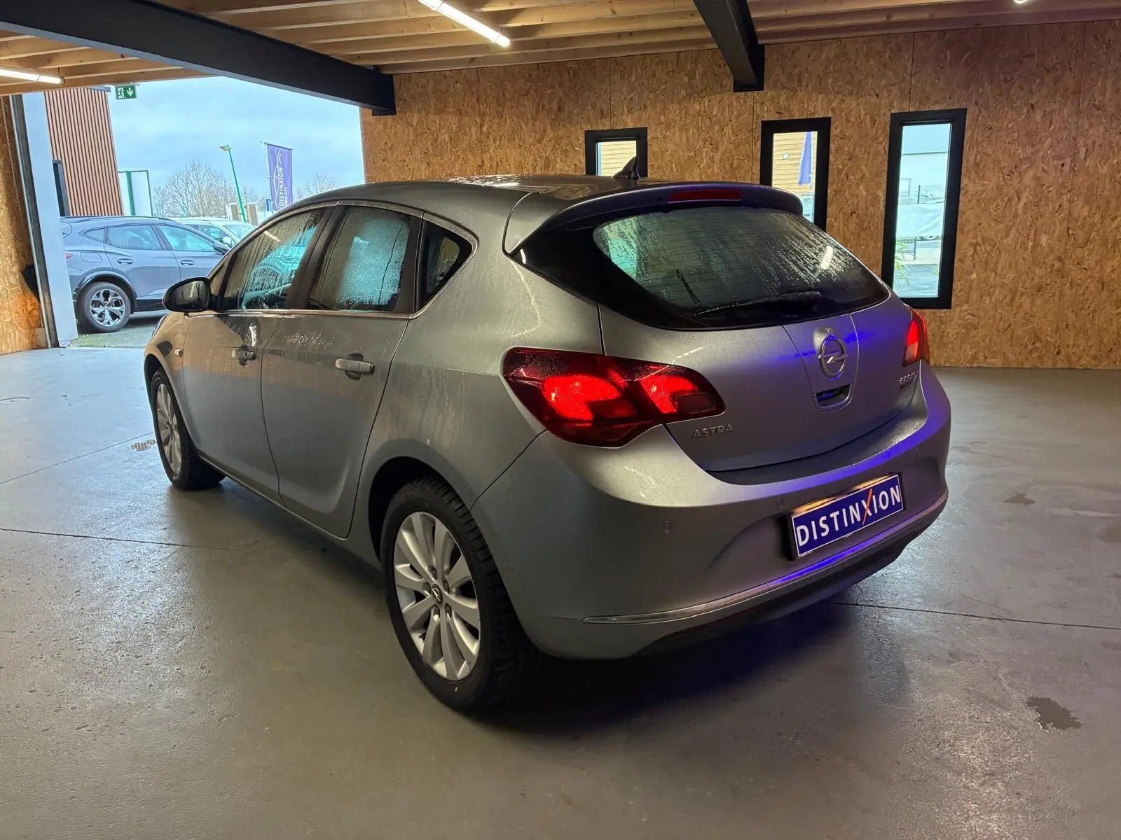 Opel Astra gris 2015 vue 3/4 arrière droit dans un garage, feux arrière allumés et plaque Distinxion visible.