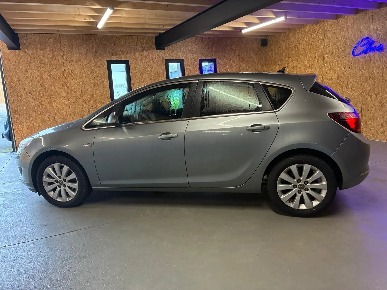 Vue latérale droite d'une Opel Astra gris métallisé de 2015, avec jantes alliage et vitres légèrement embuées.