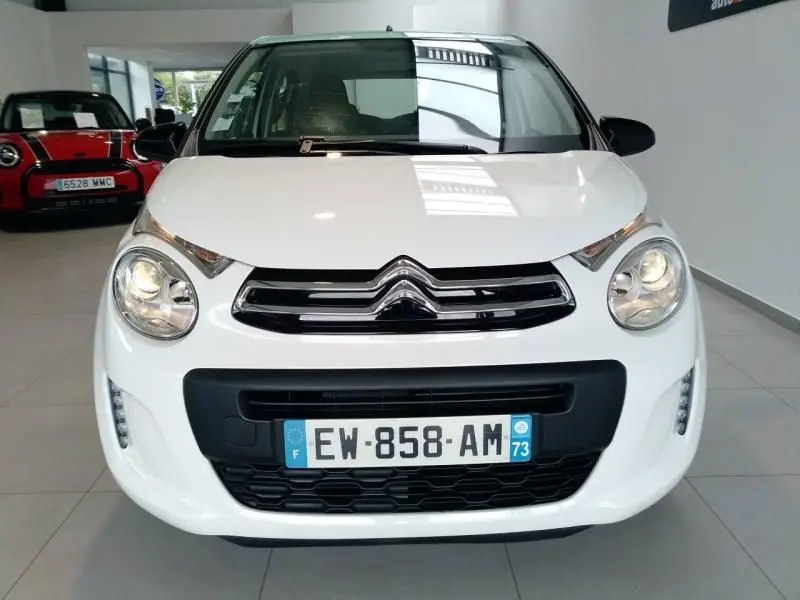 Vue frontale d'une Citroën C1 blanche 2018 avec phares allumés et calandre noire dans un showroom.