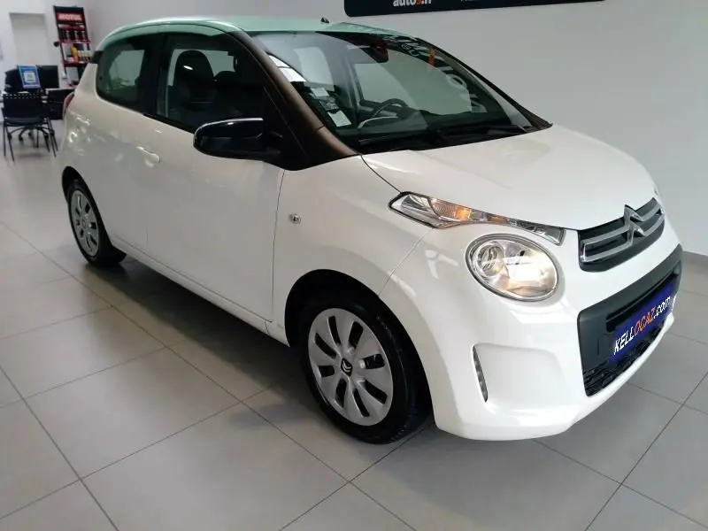 Citroën C1 blanche vue 3/4 avant droit en intérieur, avec phares allumés et rétroviseurs noirs.