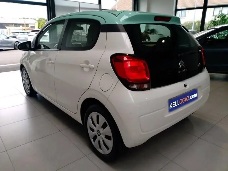 Vue 3/4 arrière droite d'une Citroën C1 blanche avec pavillon noir et feux arrière rouges dans un showroom.