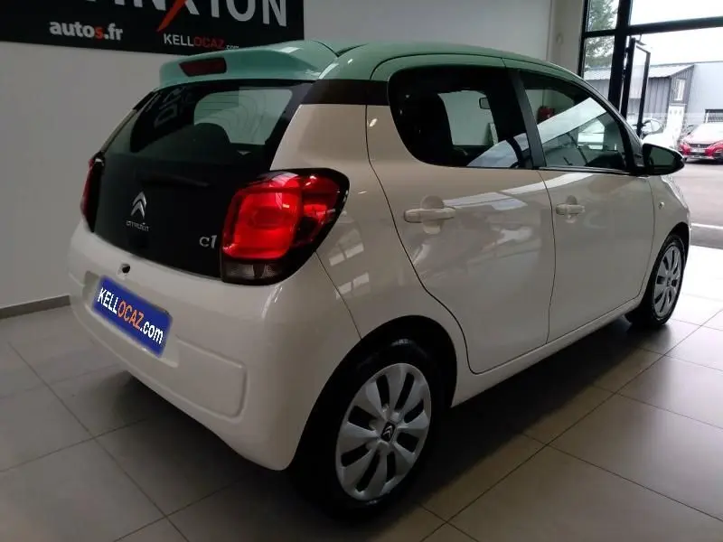 Citroën C1 blanche vue 3/4 arrière droit en intérieur, avec feux arrière rouges et enjoliveurs d'origine.