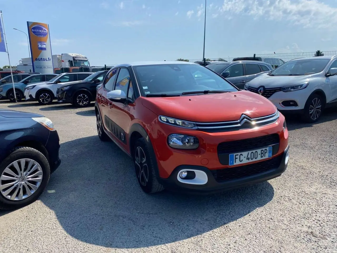 Citroën C3 orange vue 3/4 avant droit avec protections latérales noires et toit noir dans un parking.