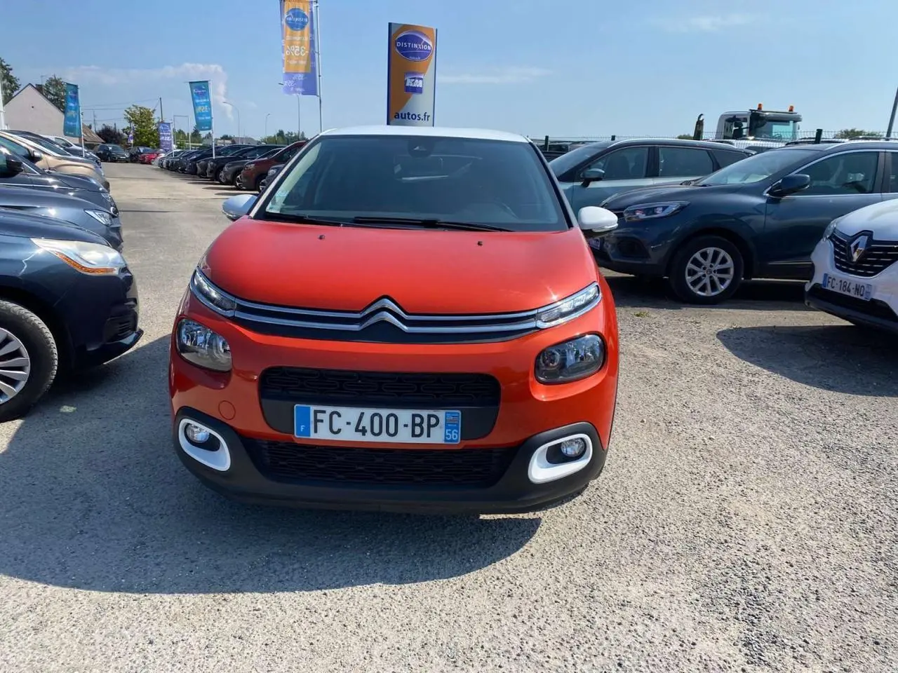 Vue de face d'une Citroën C3 orange avec toit blanc et feux avant distinctifs sur un parking extérieur.