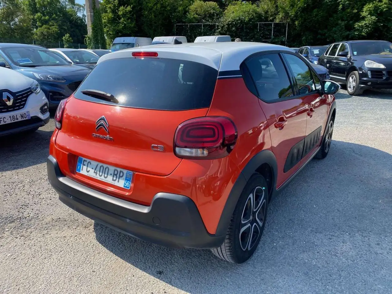 Vue 3/4 arrière droite d'une Citroën C3 orange avec toit blanc et protections latérales noires sur parking.