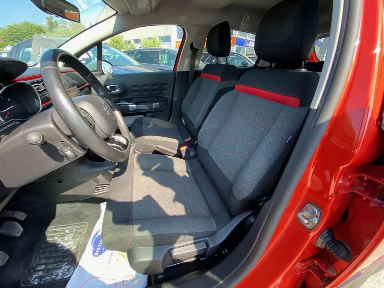 Vue intérieure côté conducteur de la Citroën C3 orange 2018, sièges tissu gris avec surpiqûres rouges et volant noir.