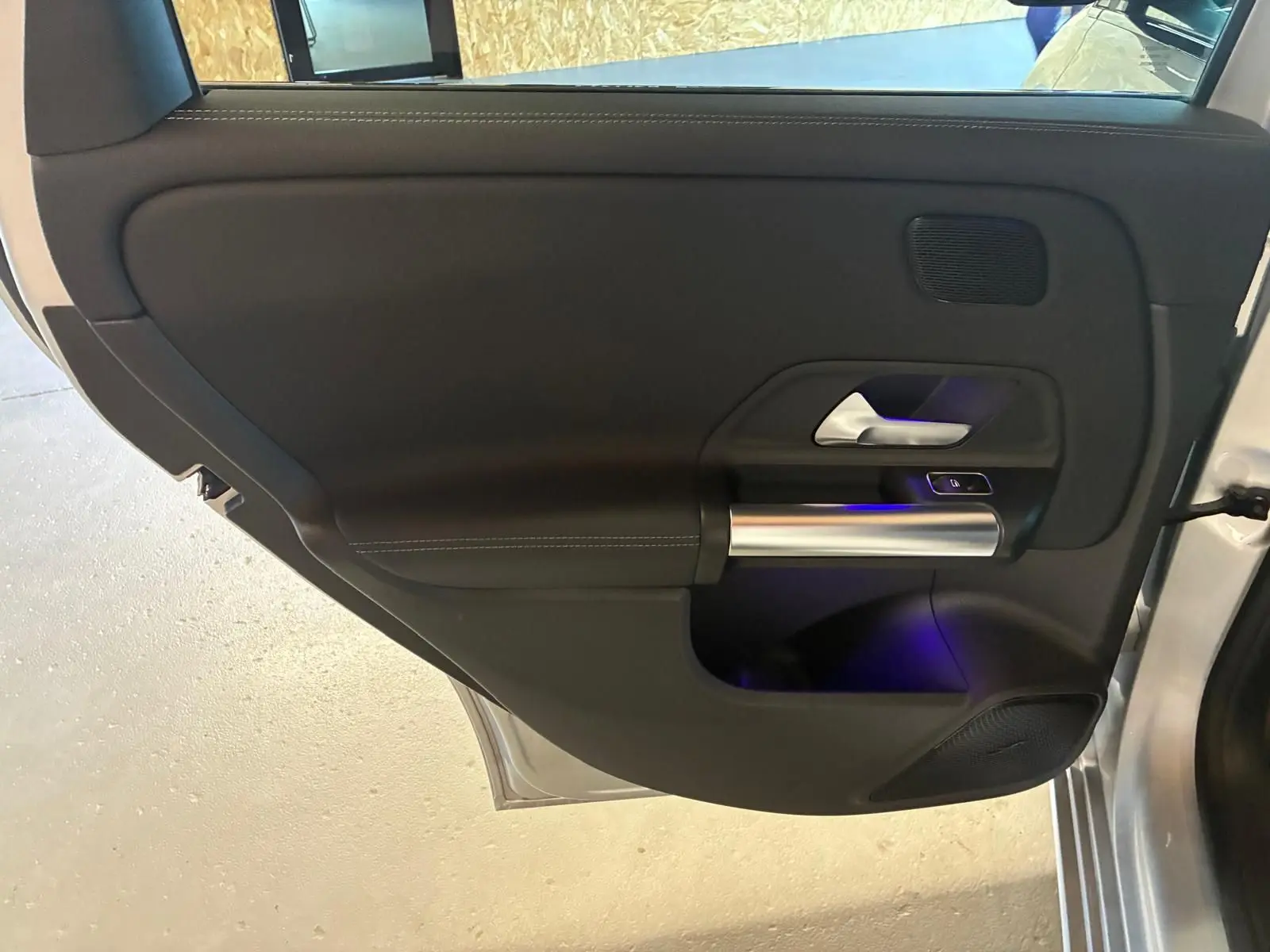 Vue intérieure de la porte arrière droite d'une Mercedes Classe B 200D gris, avec poignée alu et éclairage d'ambiance bleu.