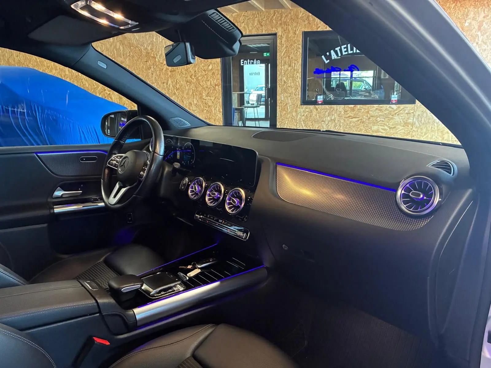 Vue intérieure côté conducteur de la Mercedes Classe B 200D gris, avec éclairage d’ambiance bleu et tableau de bord moderne.