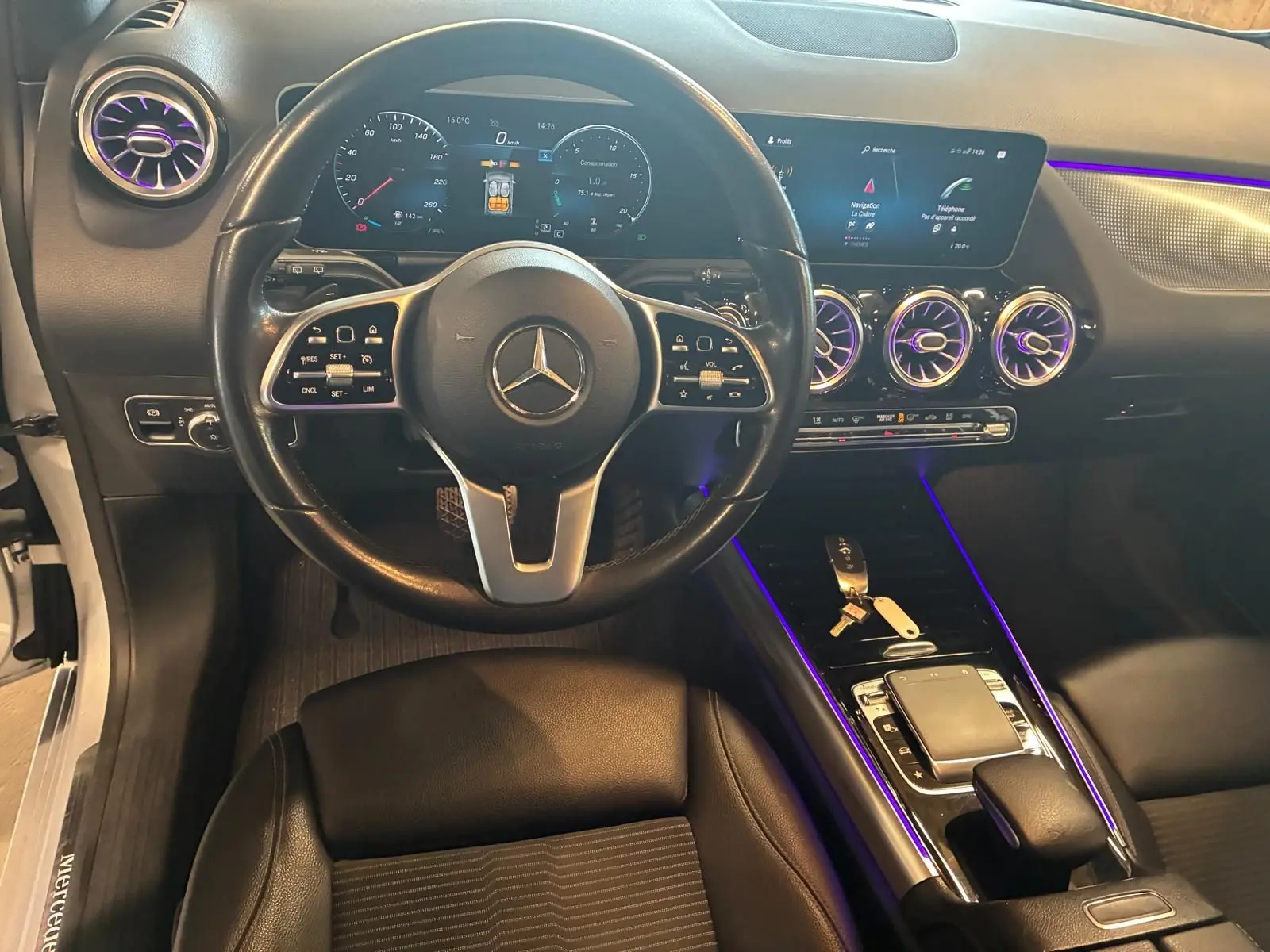 Vue intérieure centrée sur le volant et la console d'une Mercedes Classe B 200D gris, avec éclairage d'ambiance violet.