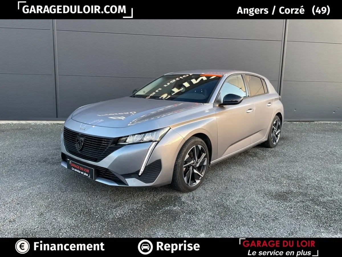 Peugeot 308 gris foncé vue 3/4 avant droit, avec calandre noire et feux LED distinctifs, stationnée devant un mur gris.