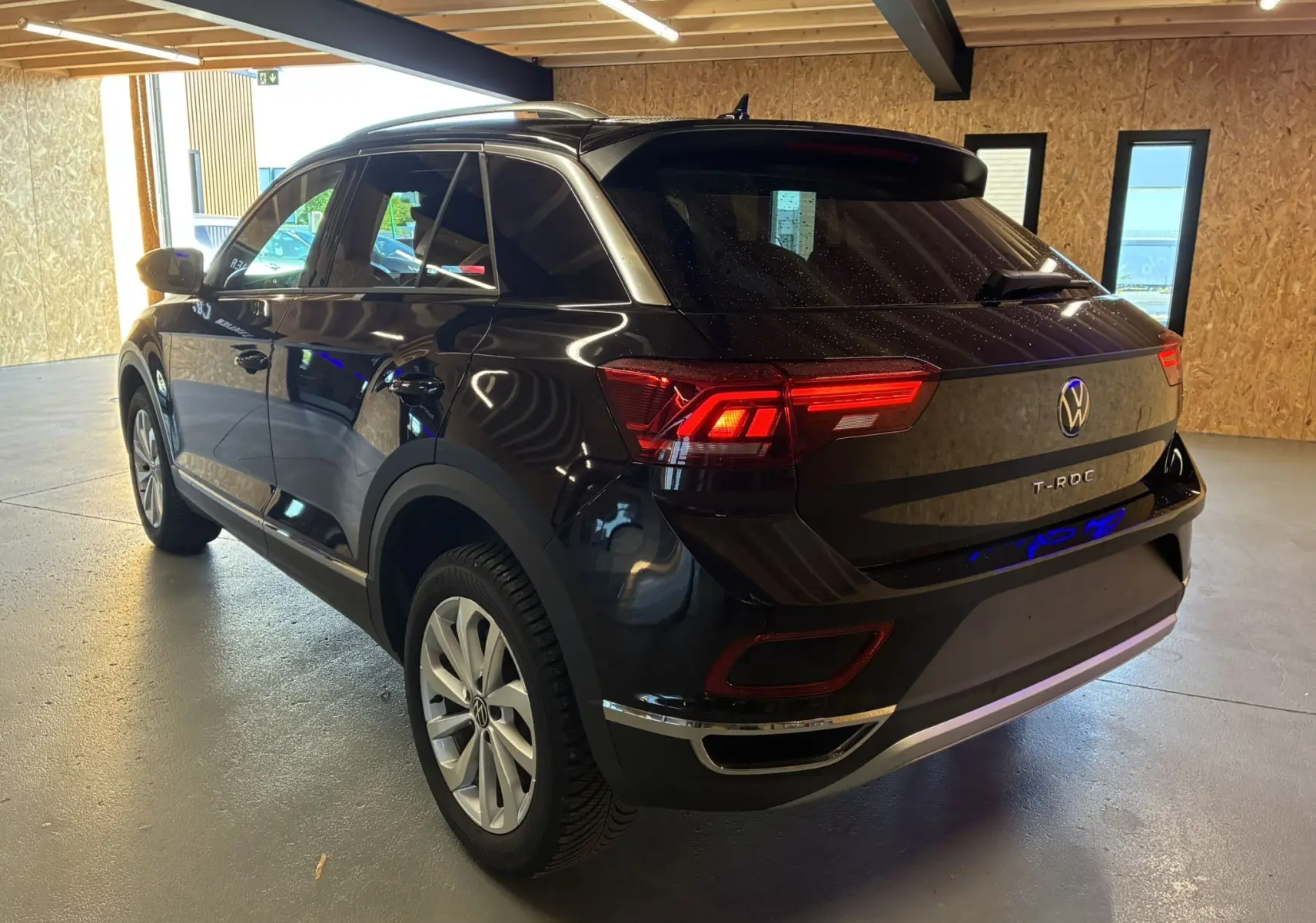 Vue 3/4 arrière droite du Volkswagen T-Roc noir 2025 avec feux arrière allumés dans un garage.