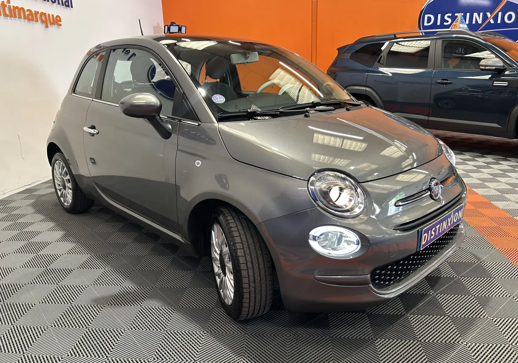 Fiat 500 gris en 3/4 avant droit, avec toit panoramique, dans un showroom au sol damier noir et gris.