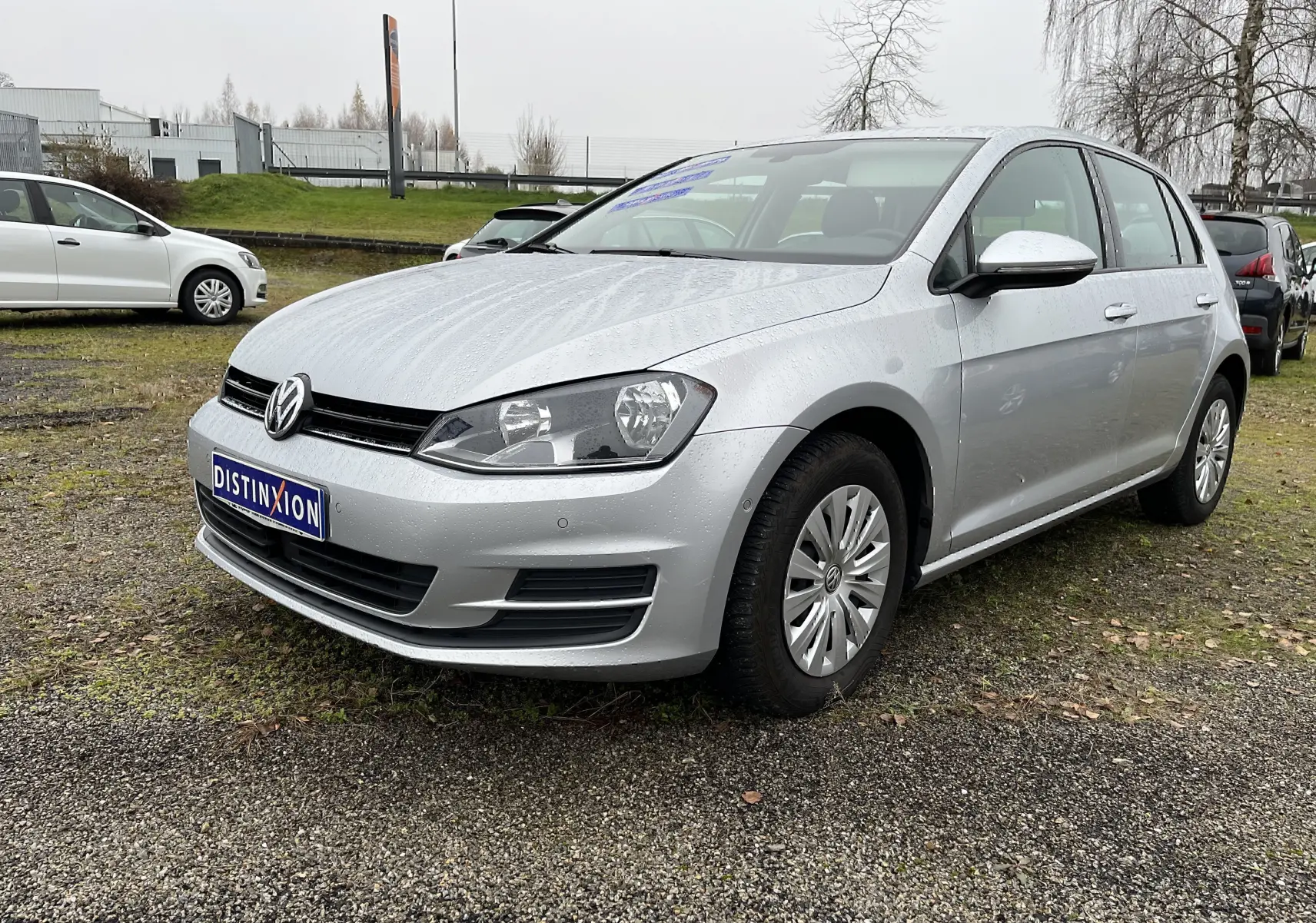 Volkswagen Golf 7 argent vue 3/4 avant droit sur parking avec carrosserie mouillée par la pluie.
