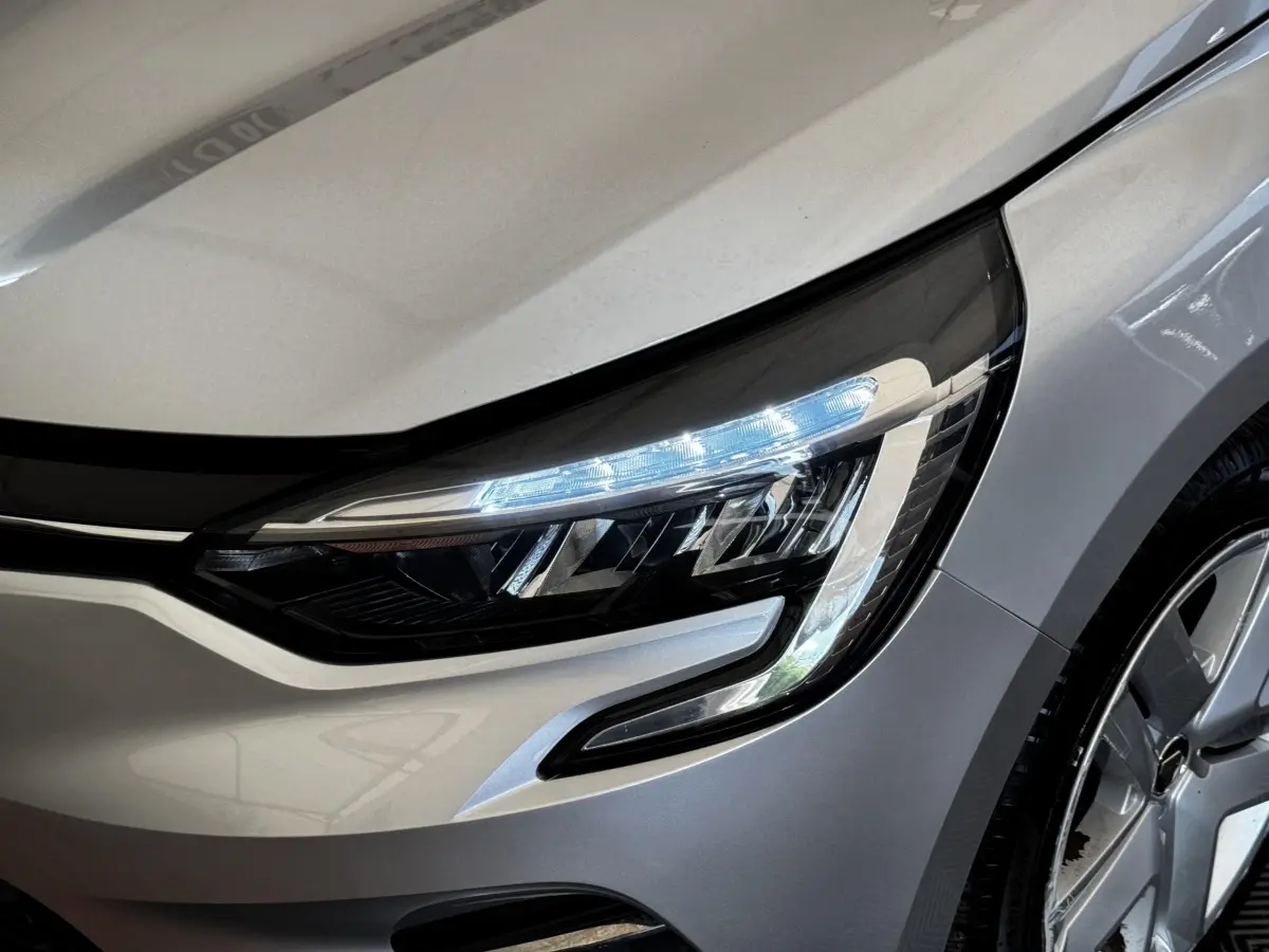 Gros plan sur le phare avant droit allumé d'une Renault Clio Business TCe 90 gris clair, avec partie de la roue visible.