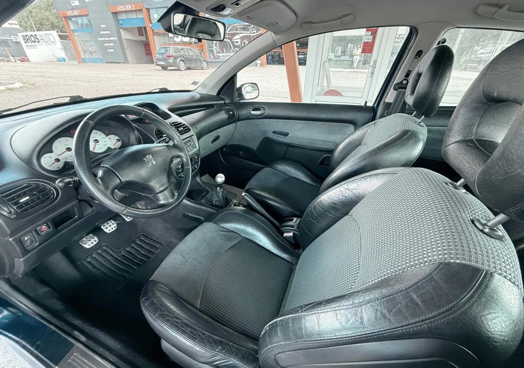 Vue intérieure côté conducteur de la Peugeot 206 bleu, montrant le volant cuir, la sellerie mixte cuir-tissu noire et la boîte manuelle.