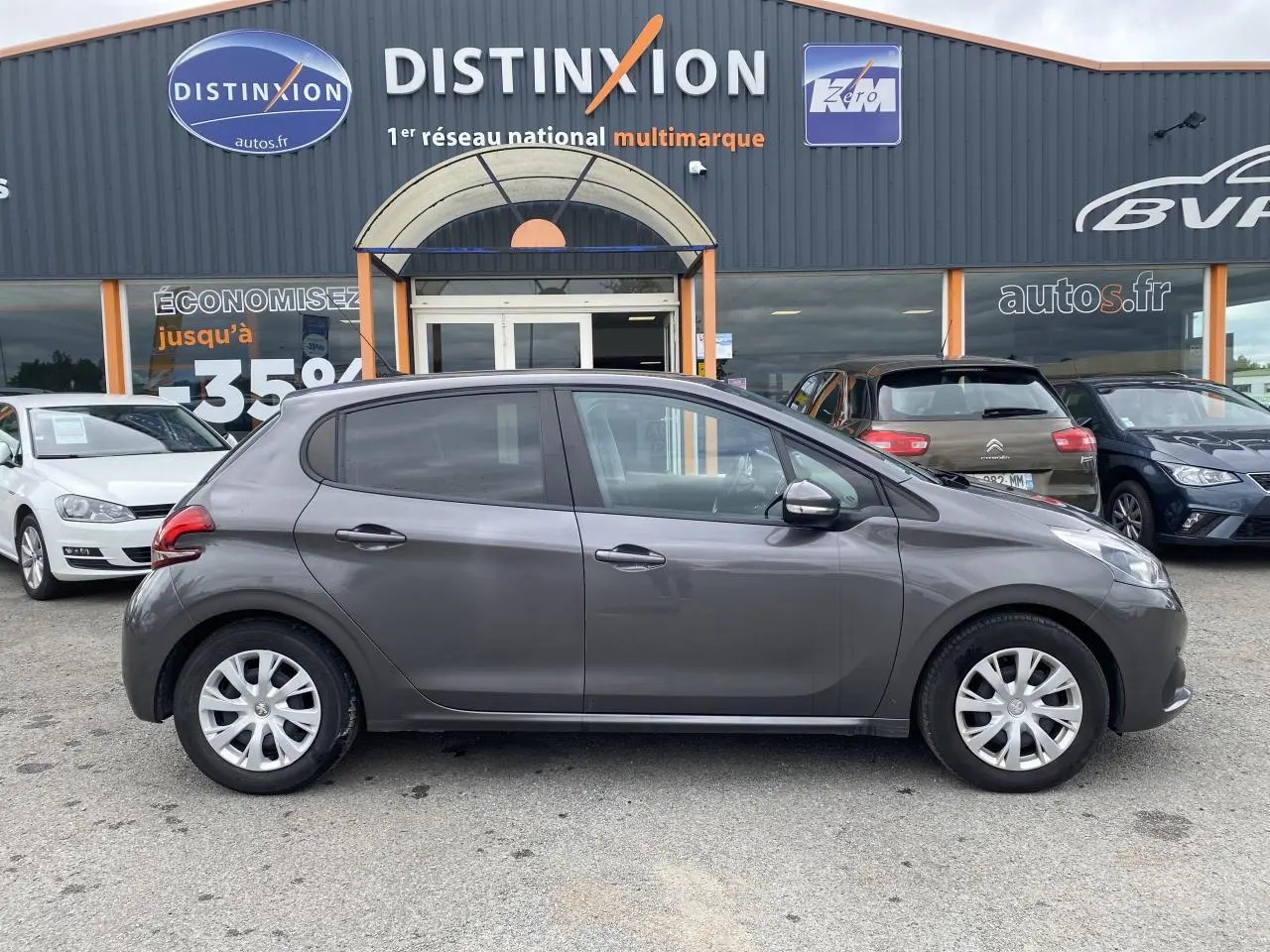 Profil côté gauche d'une Peugeot 208 gris métallisé de 2018 garée devant un concessionnaire multimarque.