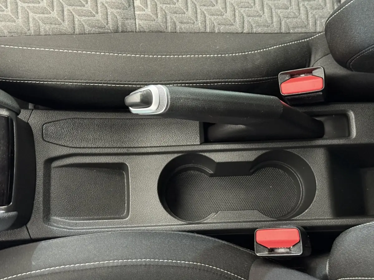 Vue plongeante sur le frein à main et porte-gobelets noirs entre les sièges avant en tissu gris de la Renault Clio blanche.