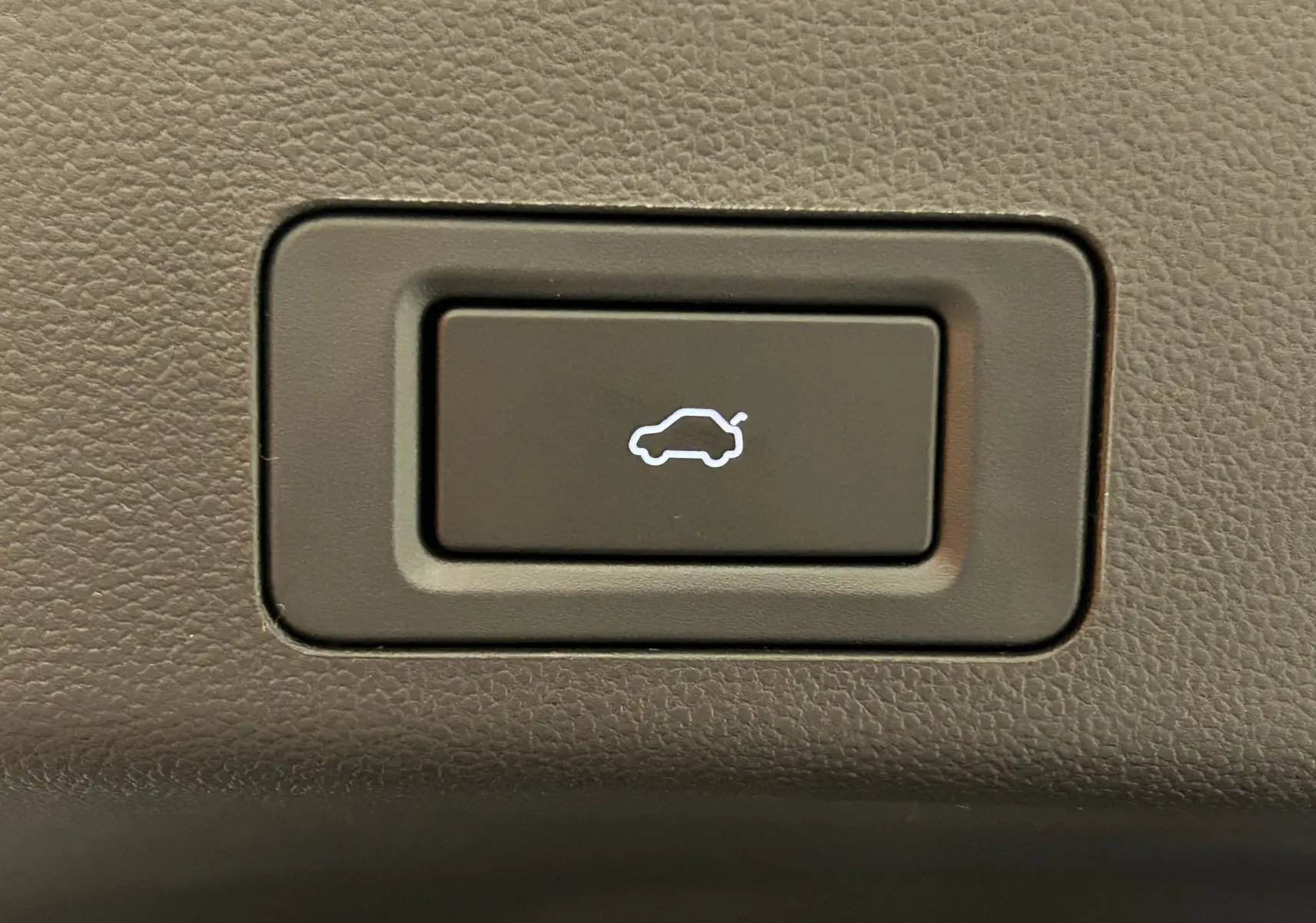 Gros plan sur le bouton d'ouverture du coffre, éclairé en blanc, sur fond de plastique texturé gris foncé.