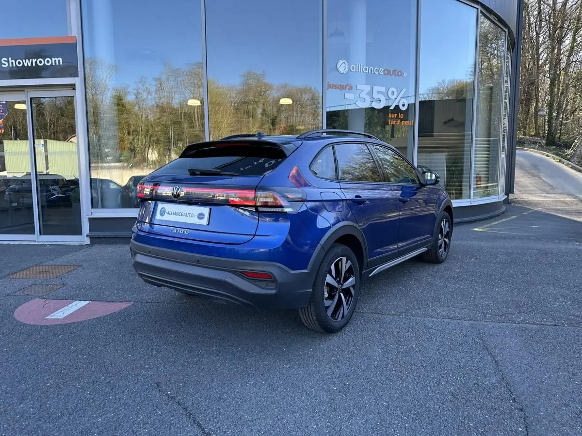 Volkswagen Taigo bleu Recife en 3/4 arrière droit, stationné devant un showroom avec vitres réfléchissantes.