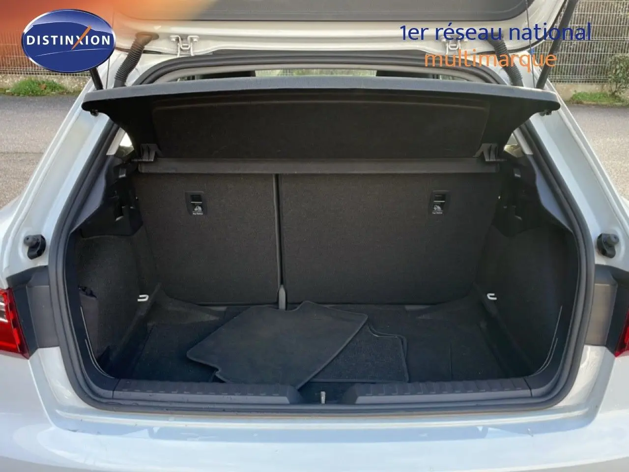 Coffre ouvert d'une Audi A1 Sportback blanc glacier métal, vue arrière montrant l'espace de chargement vide avec tapis noirs.