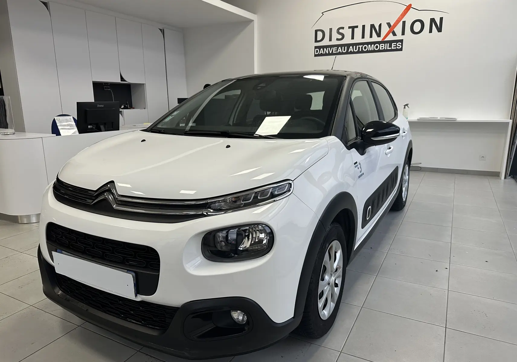 Citroën C3 blanc vue 3/4 avant droit en intérieur, avec protections latérales noires et jantes acier.