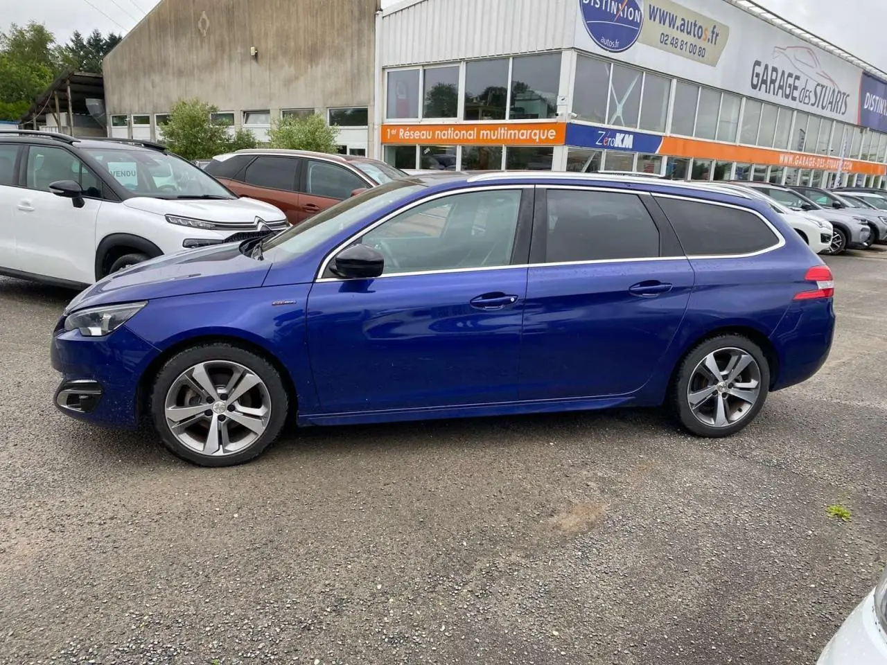 Peugeot 308 SW bleu magnétique vue de profil côté gauche sur parking devant garage multimarque.
