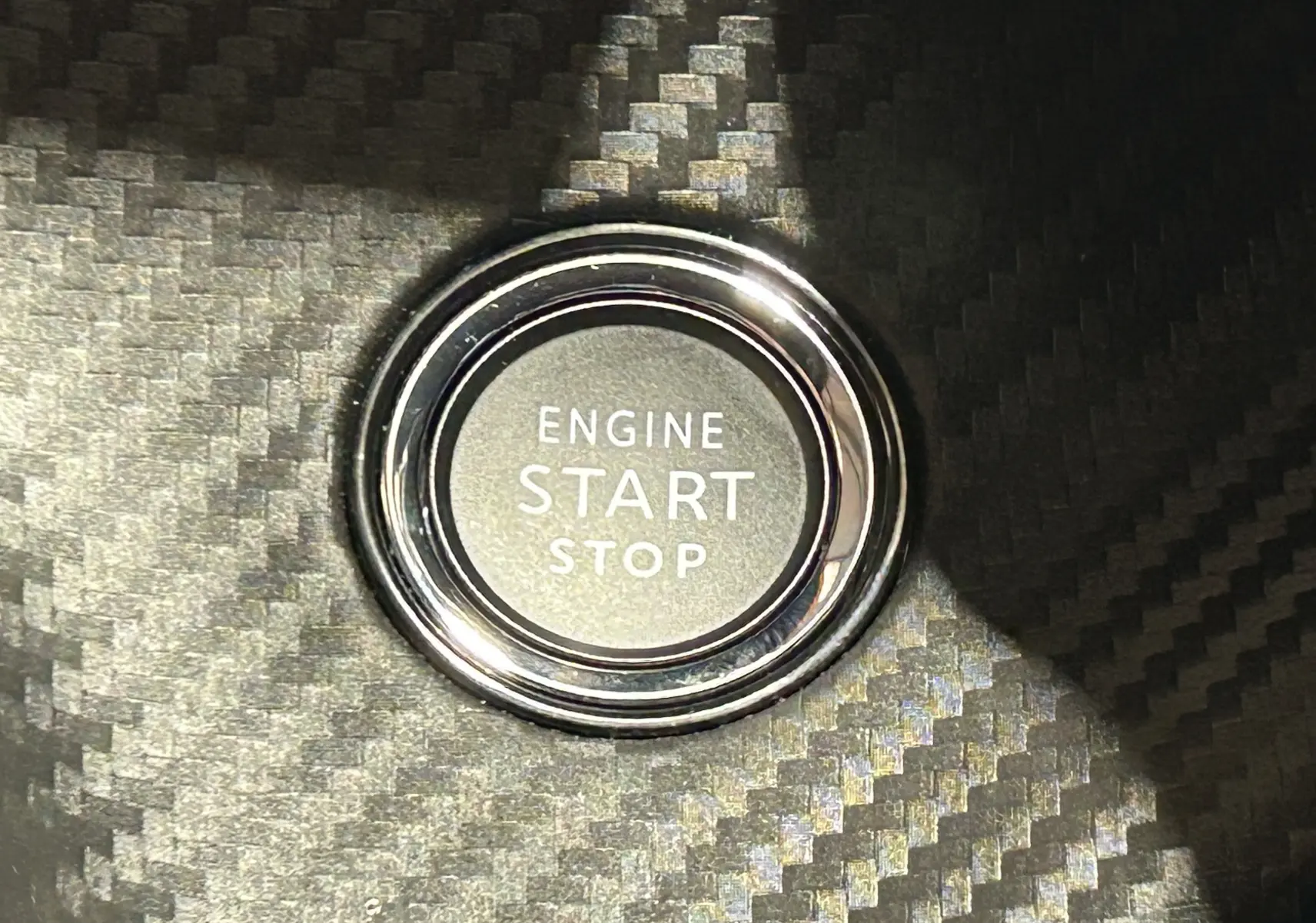 Gros plan sur le bouton Engine Start Stop entouré d’un cerclage chromé sur fond de finition carbone intérieure.