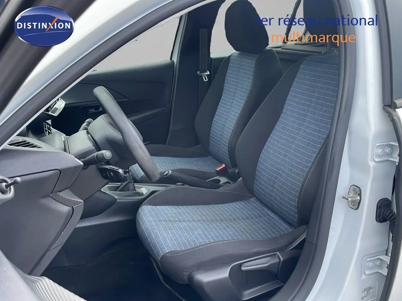 Intérieur côté gauche d'une Peugeot 208 blanc banquise 2021, sièges tissu bleu et noir, volant et levier de vitesse manuels visibles.