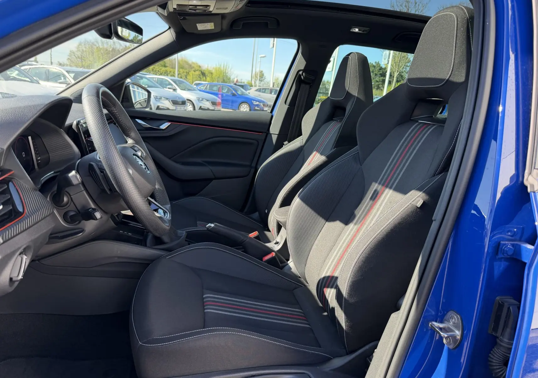 Vue intérieure côté conducteur du Skoda Kamiq bleu 2025, sièges sport noirs avec surpiqûres rouges et volant cuir trois branches.