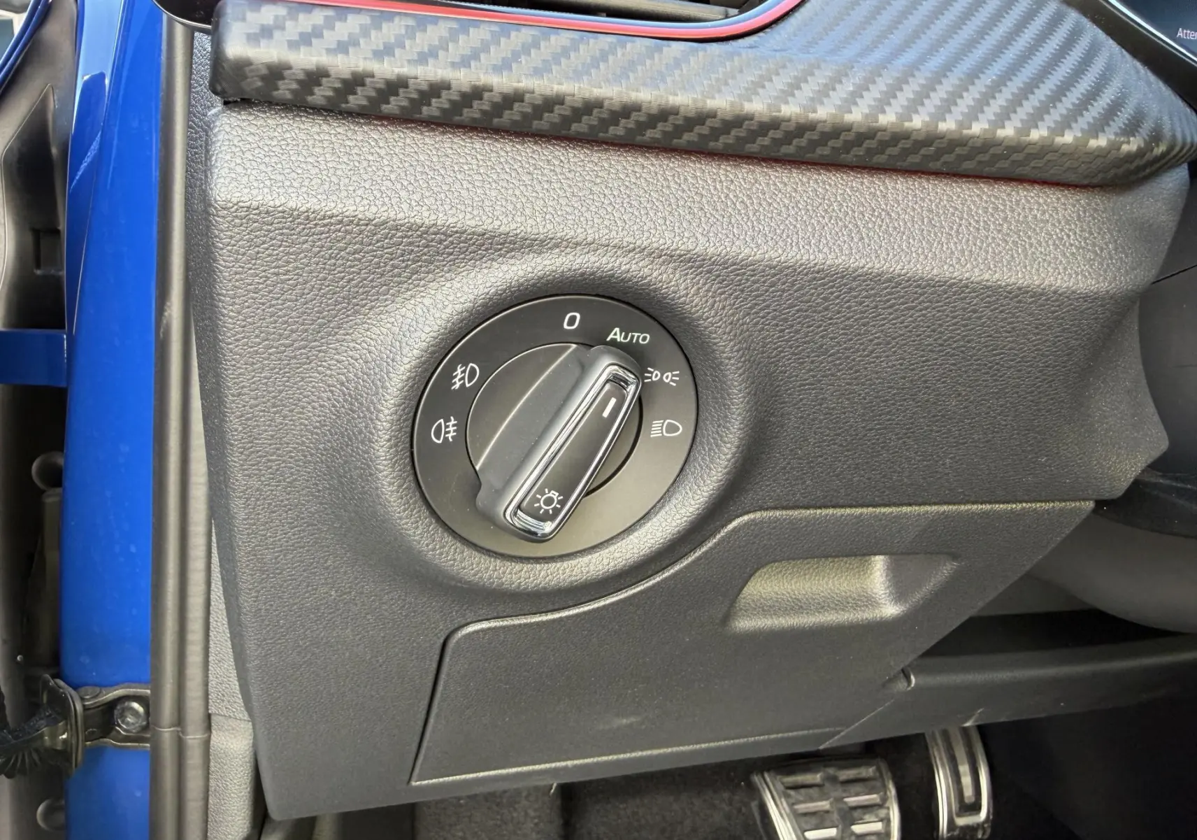 Gros plan sur le bouton de commande des phares dans l’habitacle d’une Skoda Kamiq bleu, avec finition carbone et pédales sport.