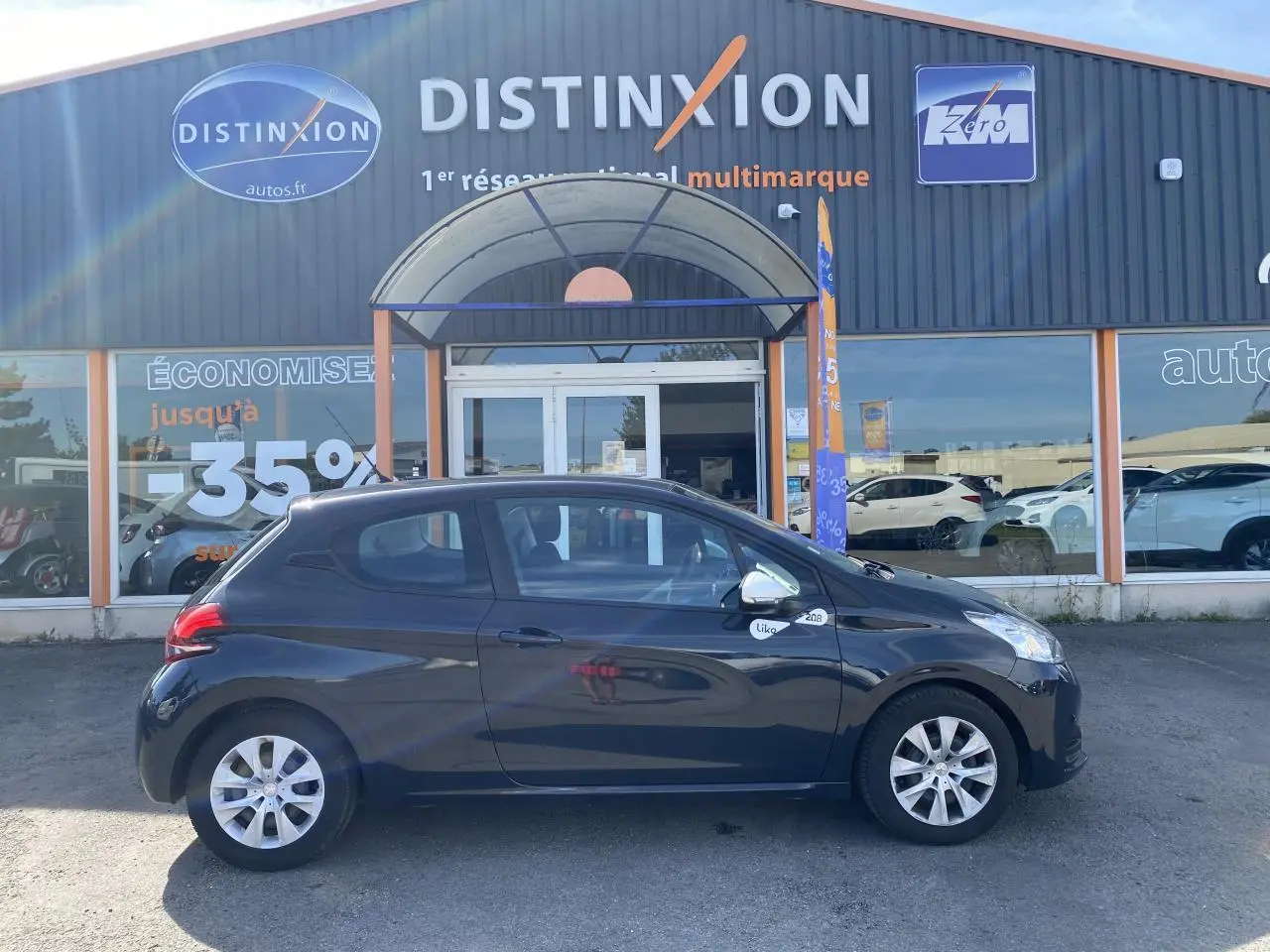 Profil côté gauche d'une Peugeot 208 gris foncé Like 2018 stationnée devant un garage multimarque.
