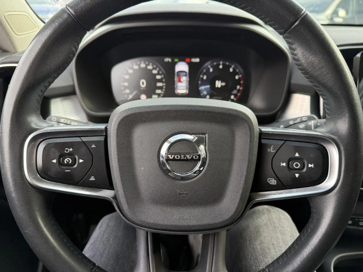 Volant cuir noir avec logo Volvo au centre, commandes intégrées, tableau de bord digital en arrière-plan.