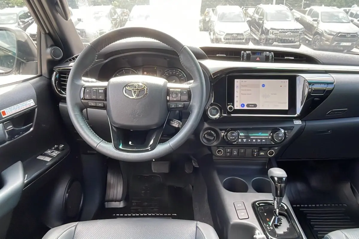 Vue intérieure frontale du tableau de bord du Toyota Hilux 4x4 blanc 2025 avec volant multifonction et écran tactile central.