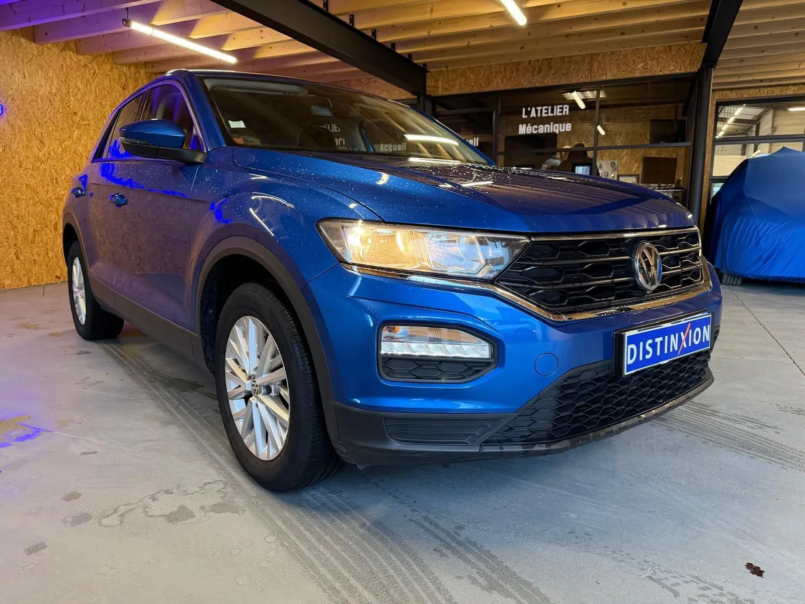 Volkswagen T-Roc bleu en 3/4 avant droit, avec calandre noire et phares allumés en intérieur d'atelier.