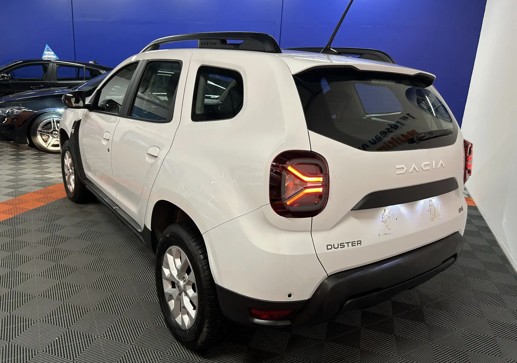 Vue 3/4 arrière droite d'un Dacia Duster blanc 2023 avec feux LED allumés et barres de toit noires.
