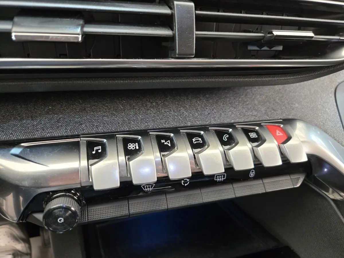 Gros plan sur la rangée de boutons argentés et noirs de la console centrale du Peugeot 5008 gris clair, avec bouton rouge d’alerte.