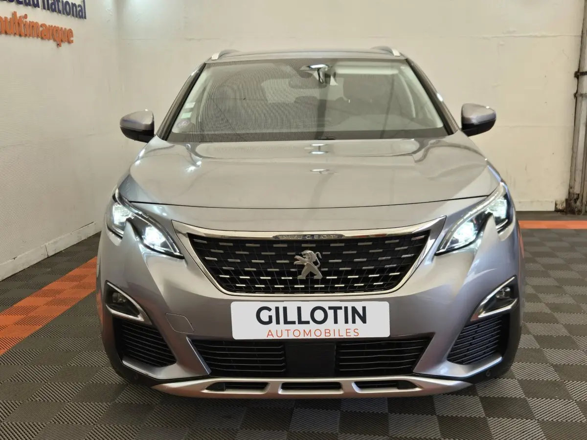 Vue frontale d'un Peugeot 5008 gris clair avec phares LED allumés et calandre chromée visible en intérieur.