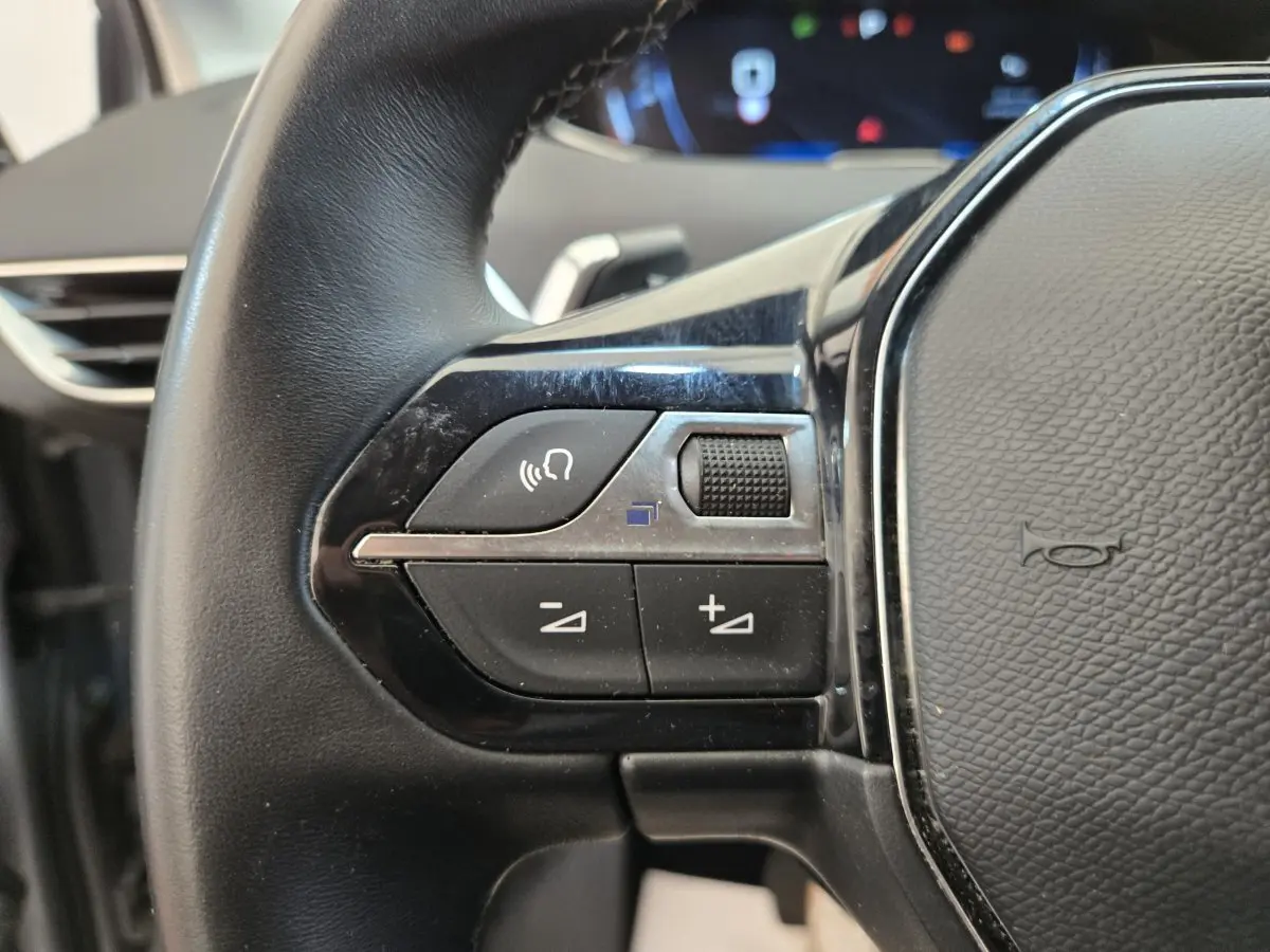 Gros plan sur les commandes gauche du volant cuir noir du Peugeot 5008 gris clair, avec boutons multifonctions et molette.