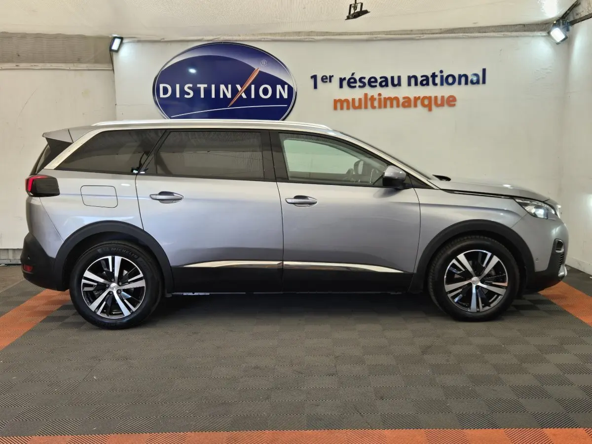 Peugeot 5008 gris clair vue de profil côté gauche, avec jantes alliage et rails de toit chromés visibles.