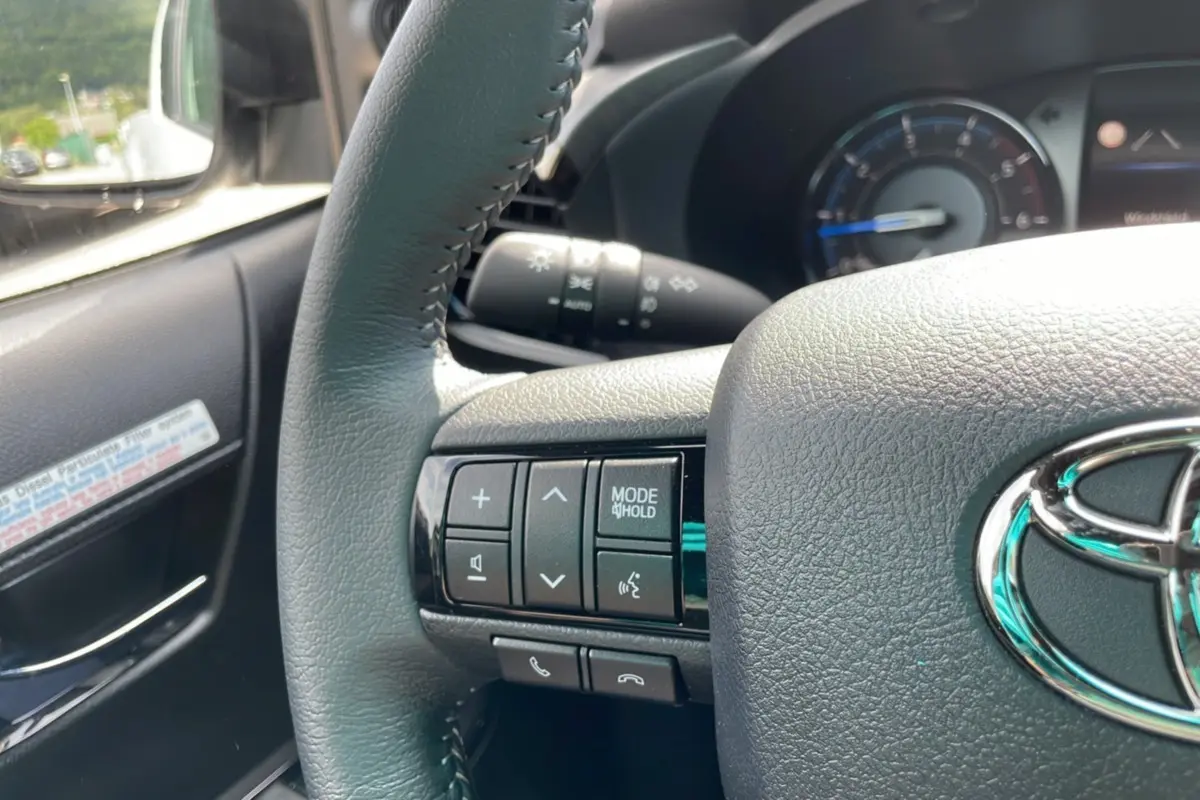 Gros plan sur le volant côté gauche du Toyota Hilux 2025 blanc, avec commandes multimédia et logo Toyota visible.